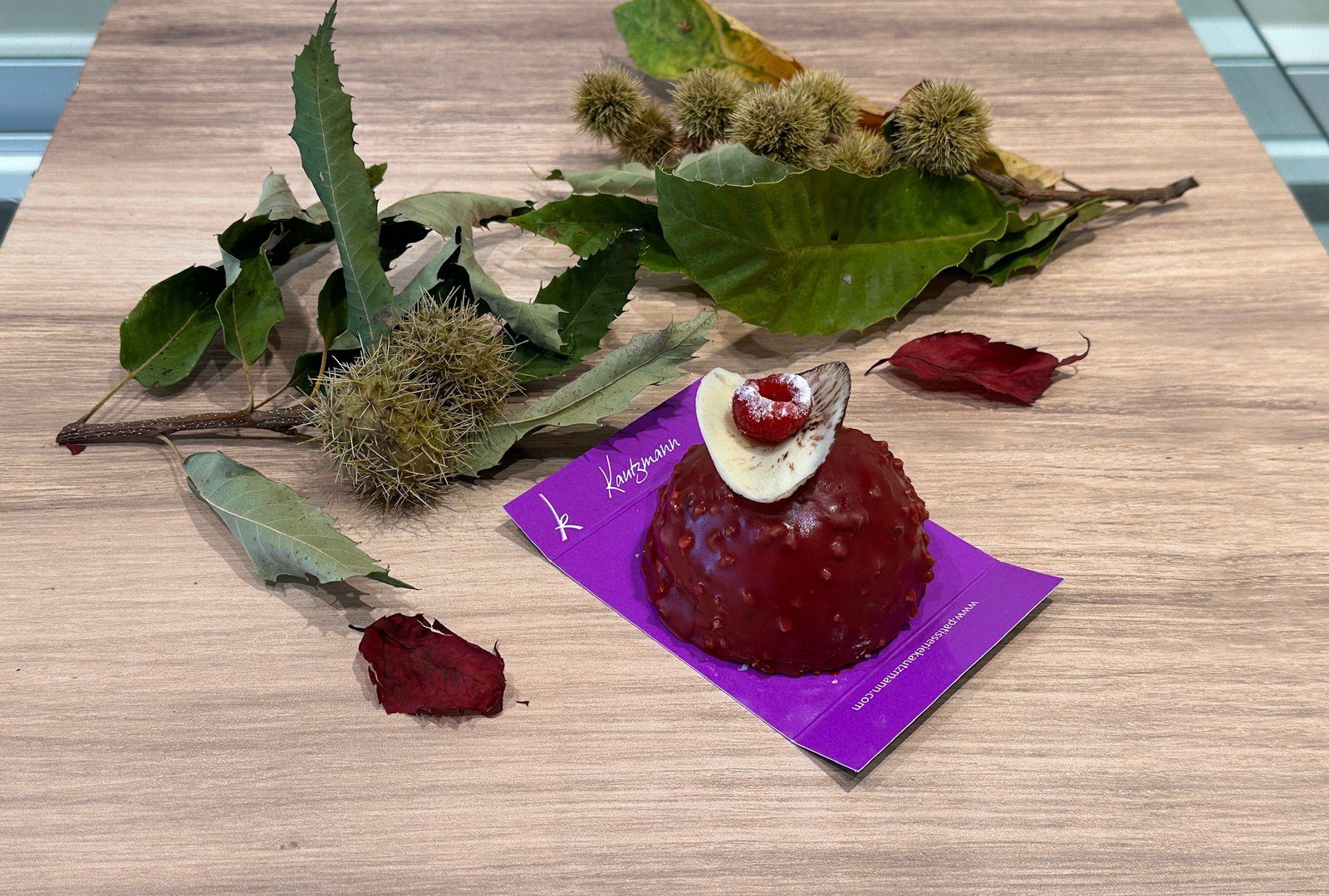 Le Rubis, dôme au glaçage rouge framboise de la Pâtisserie Kautzmann, posé sur un carton violet près de bogues de châtaignier